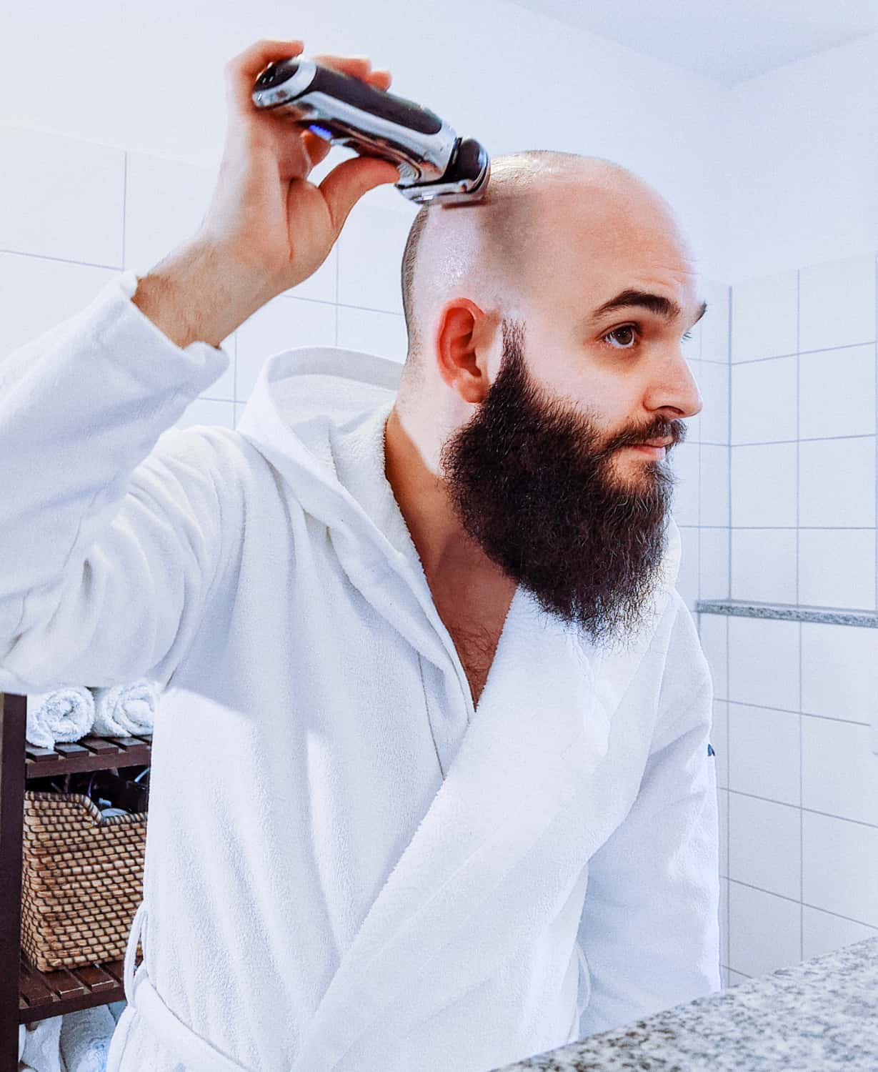 Ein Mann im Bademantel rasiert sich täglich die Glatze mit einem Elektrorasierer im Badezimmer, während er in den Spiegel schaut.