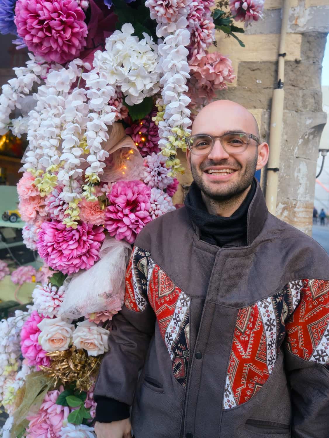 Ein Mann mit Glatze und Brille steht lächelnd vor einem üppigen Blumenarrangement aus weißen, rosa und lila Blüten. Er trägt eine Jacke mit design Mustern und genießt den Moment.