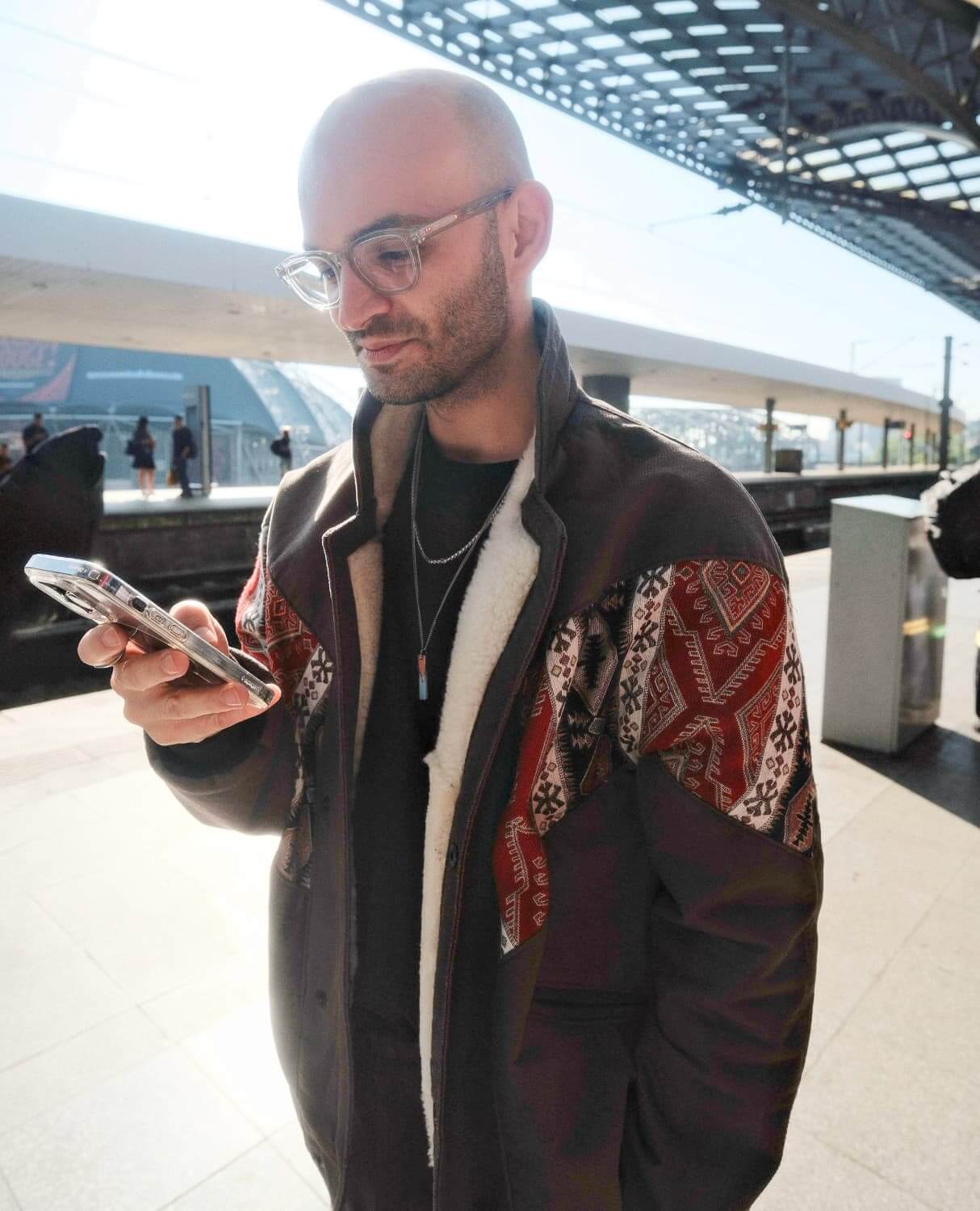 Ein Mann mit Glatze und Brille steht auf einem Bahnsteig und schaut auf sein Smartphone. Er trägt eine Jacke mit design Mustern und genießt den Tag.