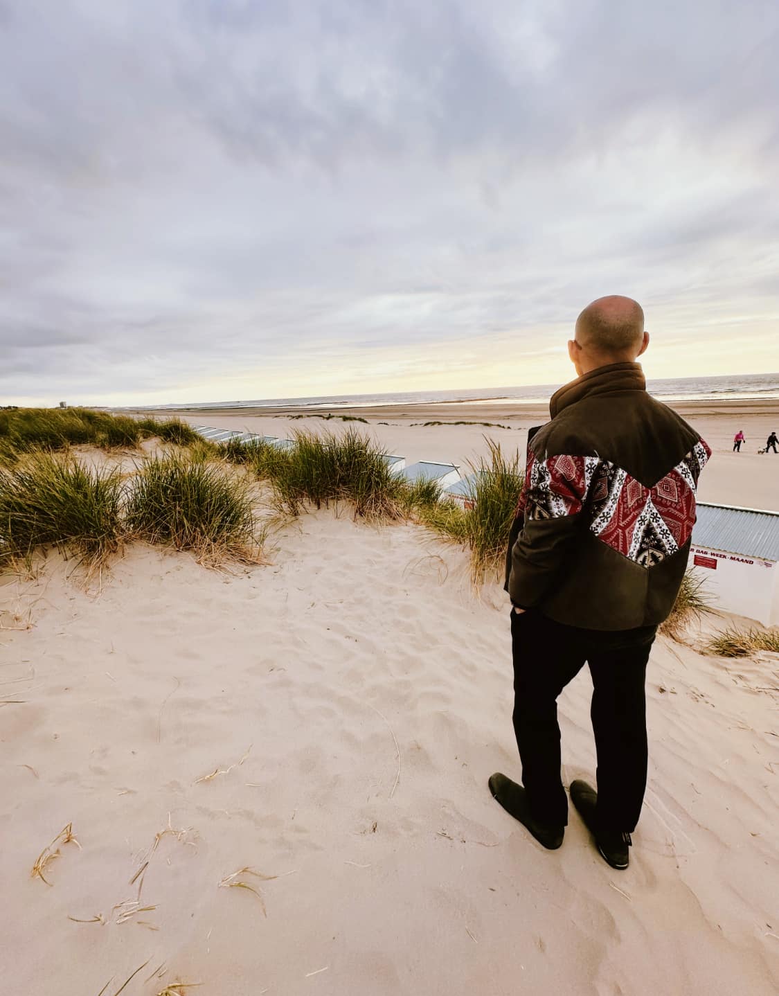 Eine Person mit Glatze steht am Strand und blickt in die Ferne. Sie trägt eine Jacke mit Design Mustern. Das Bild zeigt das Thema "Leben mit Glatze" und strahlt Ruhe und Reflexion aus.