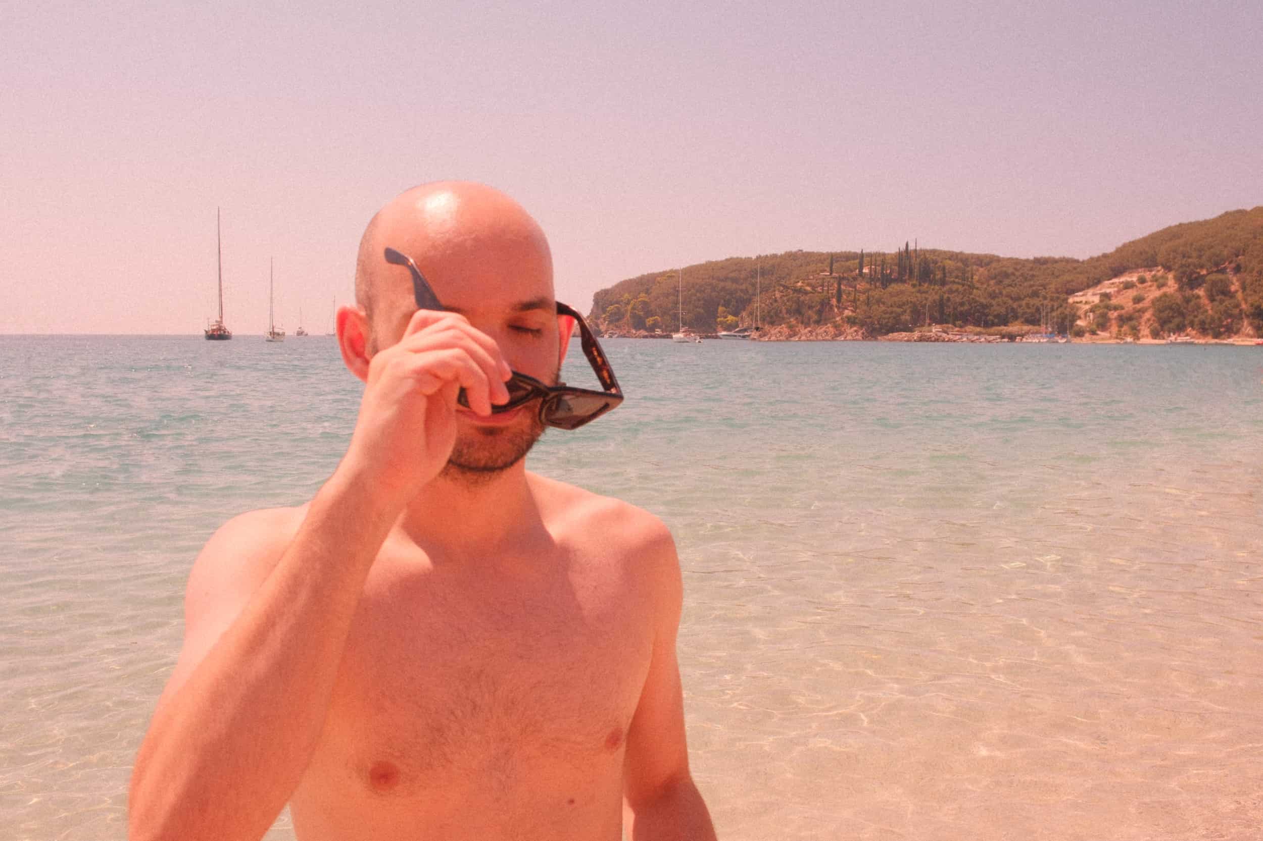 Mann mit Glatze setzt am Strand Sonnenbrille auf. Meer und Boote im Hintergrund. Perfekte Szene zur Demonstration von Kopfhaut-Sonnenschutz.