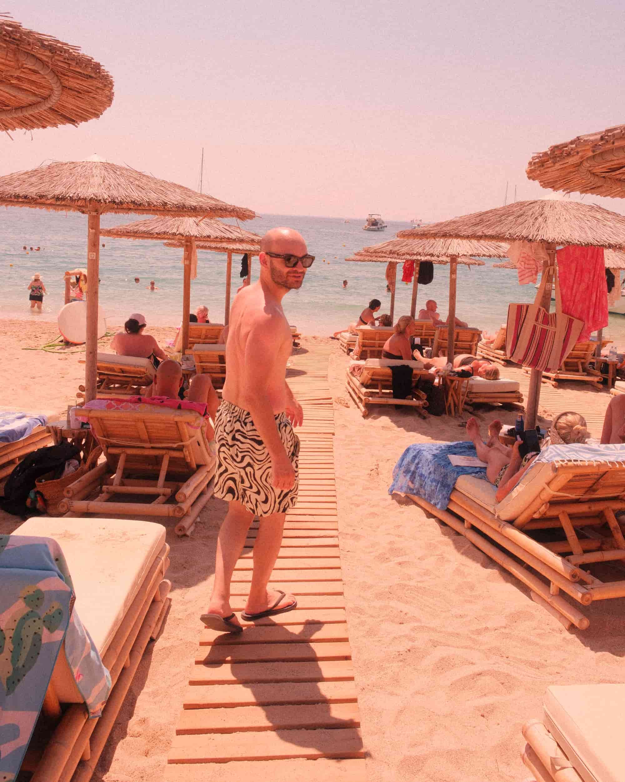 Mann mit Glatze am Strand, umgeben von Sonnenschirmen und Liegestühlen. Sonniger Tag, Meer im Hintergrund. Perfekte Szene für Kopfhaut-Sonnenschutz.