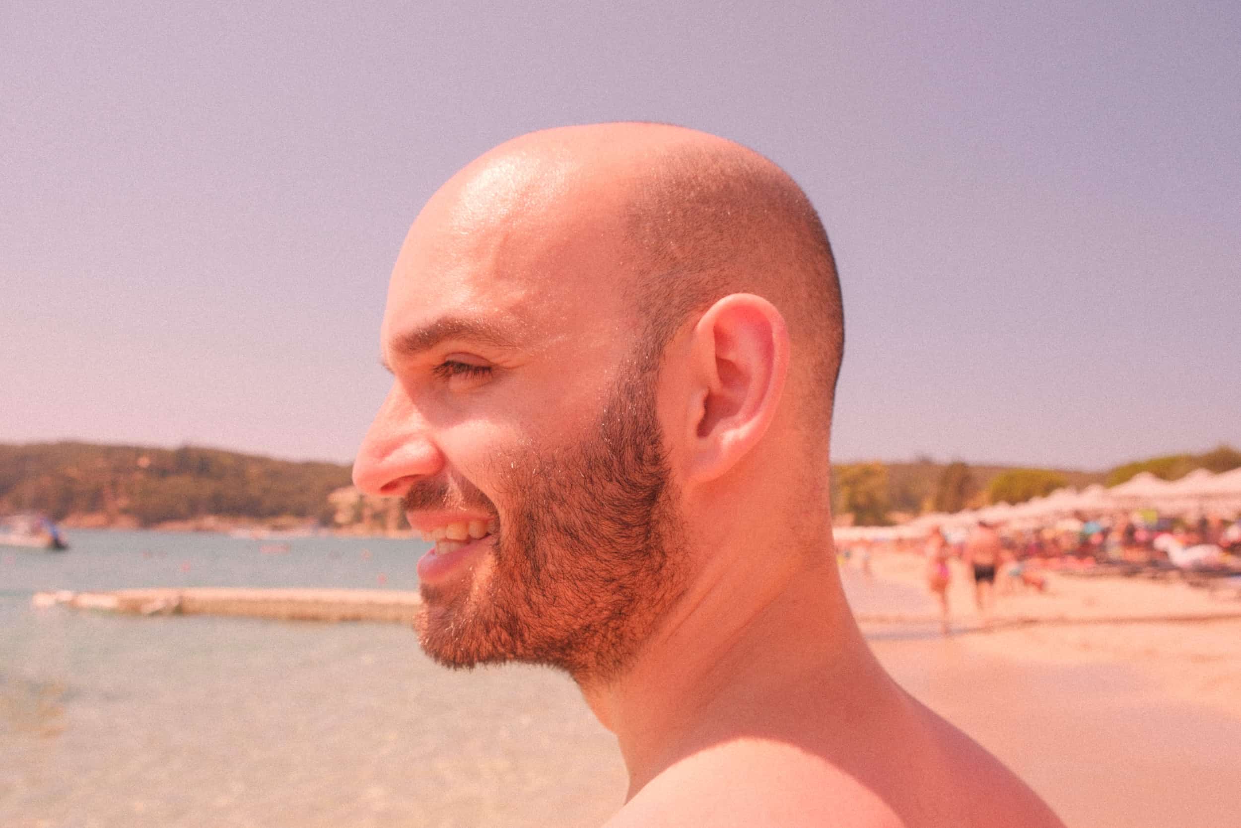 Profilansicht eines glücklichen Mannes mit Glatze am sonnigen Strand, breites Lächeln, gebräunte Haut, Meer und Strandbesucher im Hintergrund