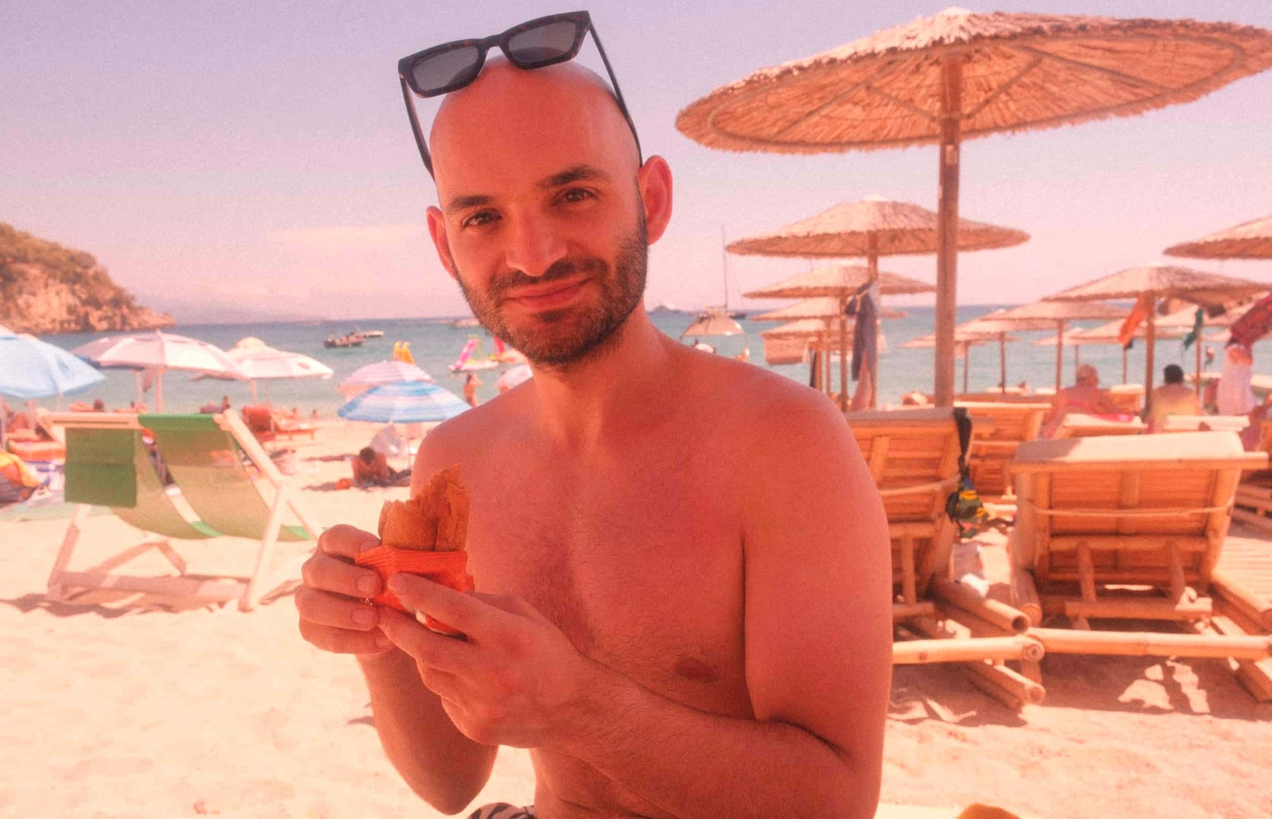 Selbstbewusster Mann mit Glatze genießt Strand-Urlaub, Sonnenbrille auf dem Kopf, lächelnd mit Snack in der Hand, Sonnenschirme im Hintergrund