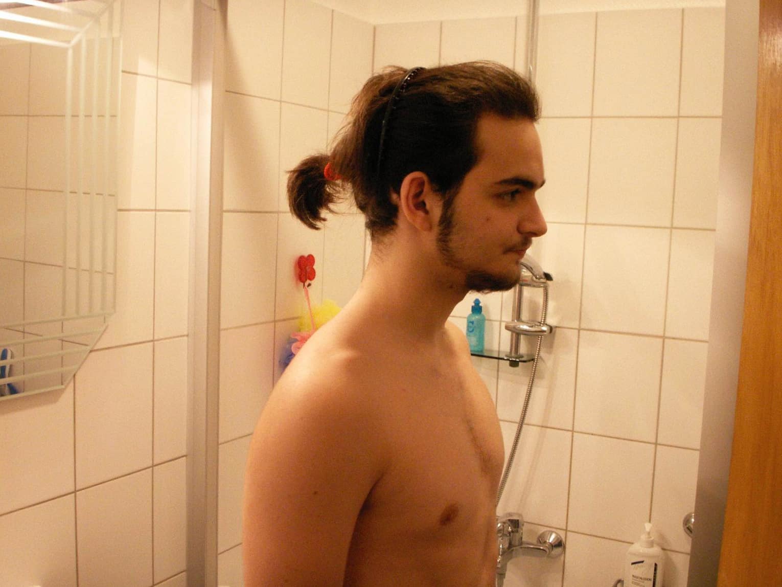 Mann mit Dutt im Badezimmer, symbolisiert humorvoll die Verzweiflung bei Haarausfall und den Versuch, Hausmittel anzuwenden.