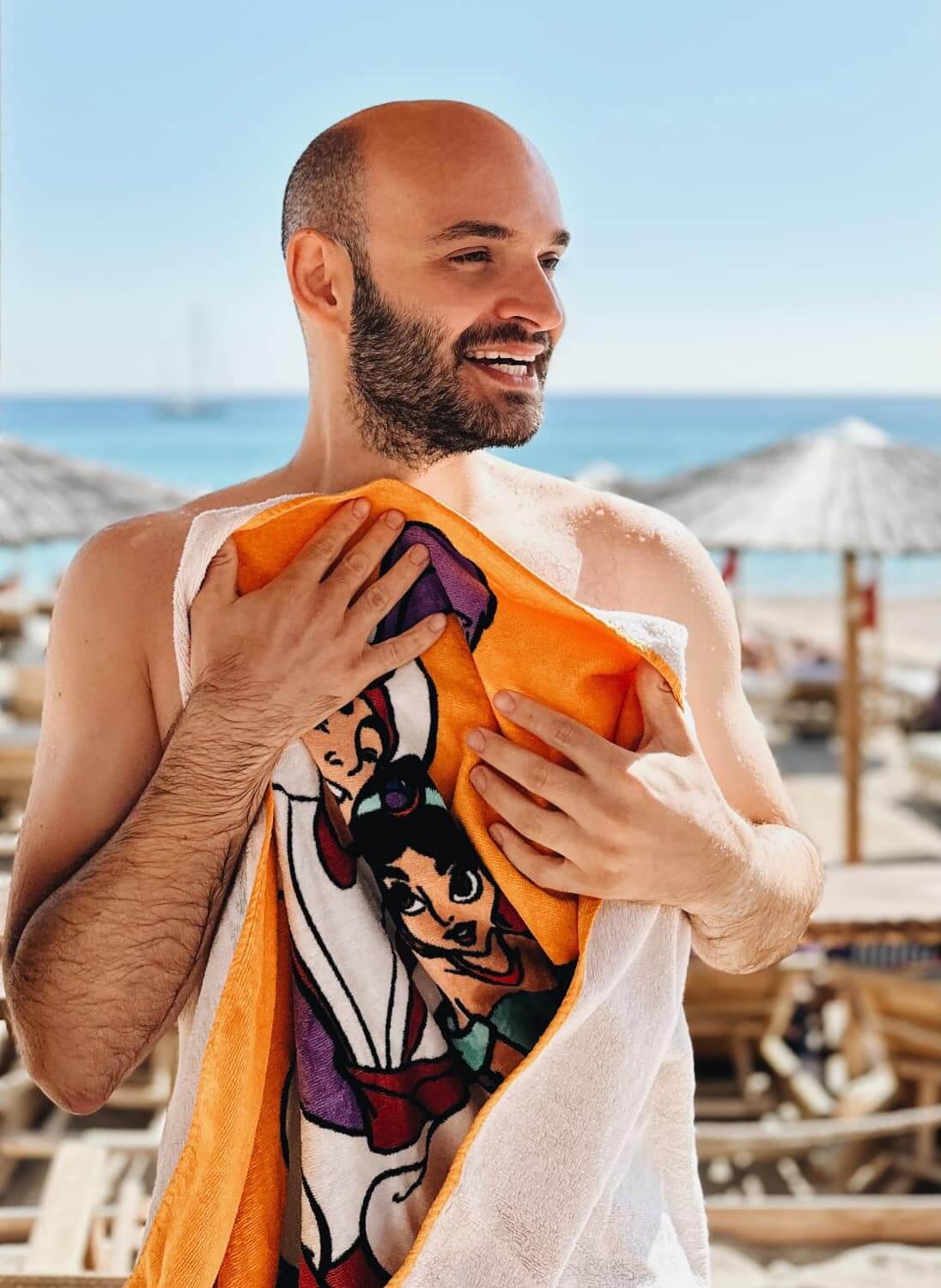 Selbstbewusster Mann mit Glatze am Strand, orangefarbenes Handtuch mit Comic-Figur haltend, strahlendes Lächeln