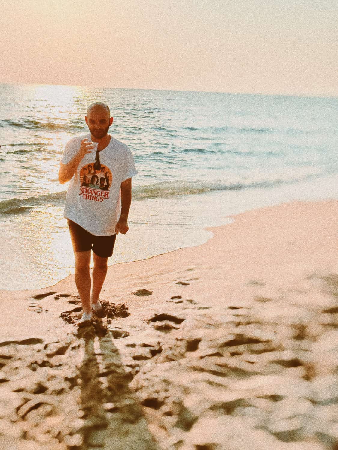 Mann mit Glatze am Strand bei Sonnenuntergang, trägt Stranger Things T-Shirt, entspannte Haltung, Fußspuren im Sand