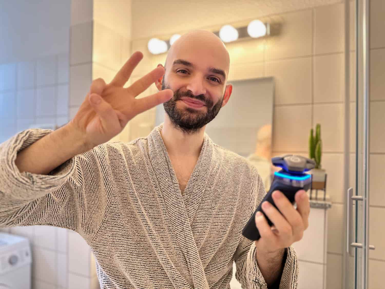 Glücklicher Mann mit Glatze macht Selfie im Badezimmer, zeigt Victory-Zeichen und hält elektrischen Rasierer - Selbstbewusstsein trotz Haarausfall