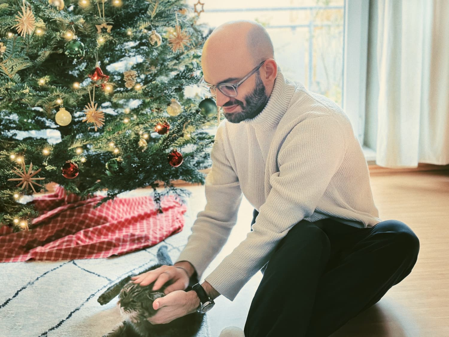 Mann im weißen Rollkragenpullover streichelt eine Katze vor geschmücktem Weihnachtsbaum, gemütliche Wohnzimmeratmosphäre