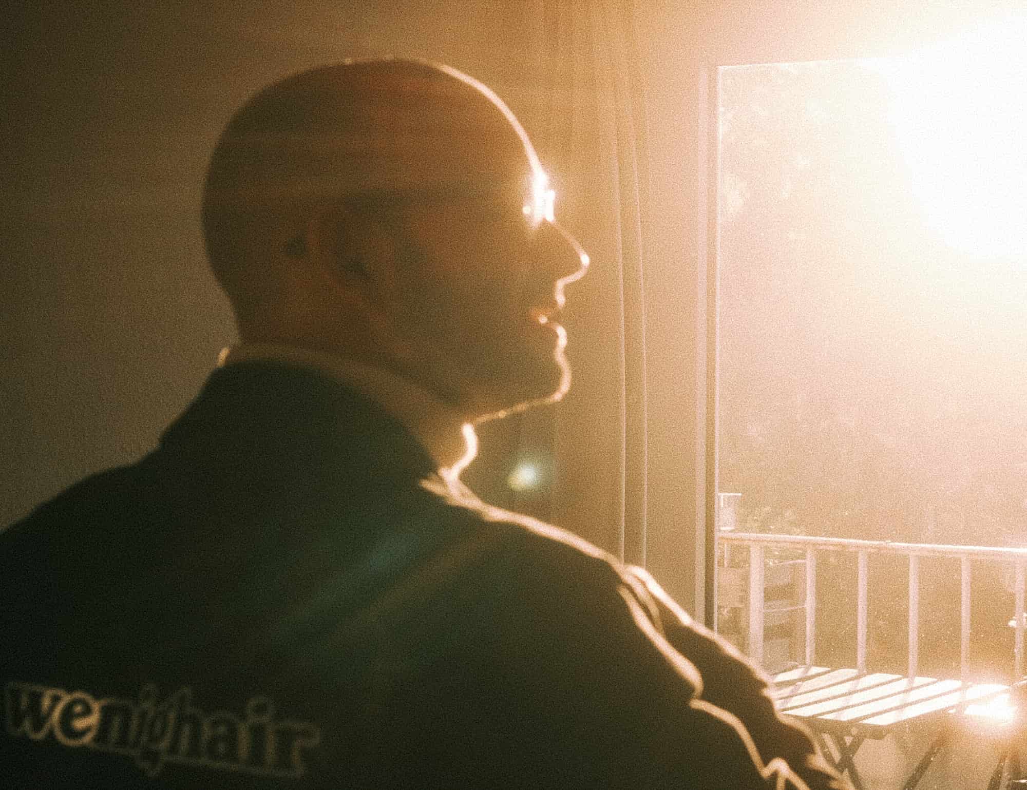 Atmosphärische Silhouette eines Mannes mit Glatze im Gegenlicht des Sonnenuntergangs auf dem Balkon, wenighair-Branding sichtbar