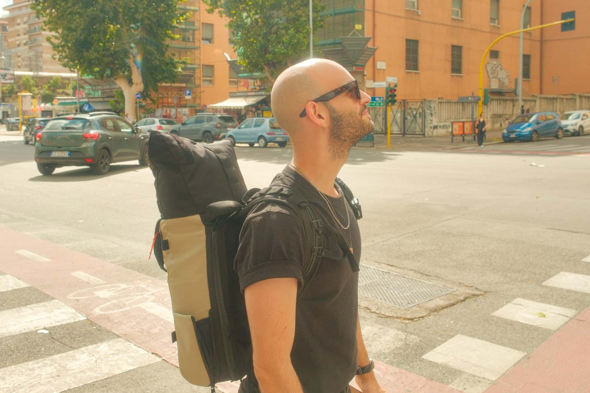 Junger Mann mit Glatze in schwarzem T-Shirt trägt einen beigen Reiserucksack auf einer italienischen Straße, urbanes Lifestyle-Motiv.