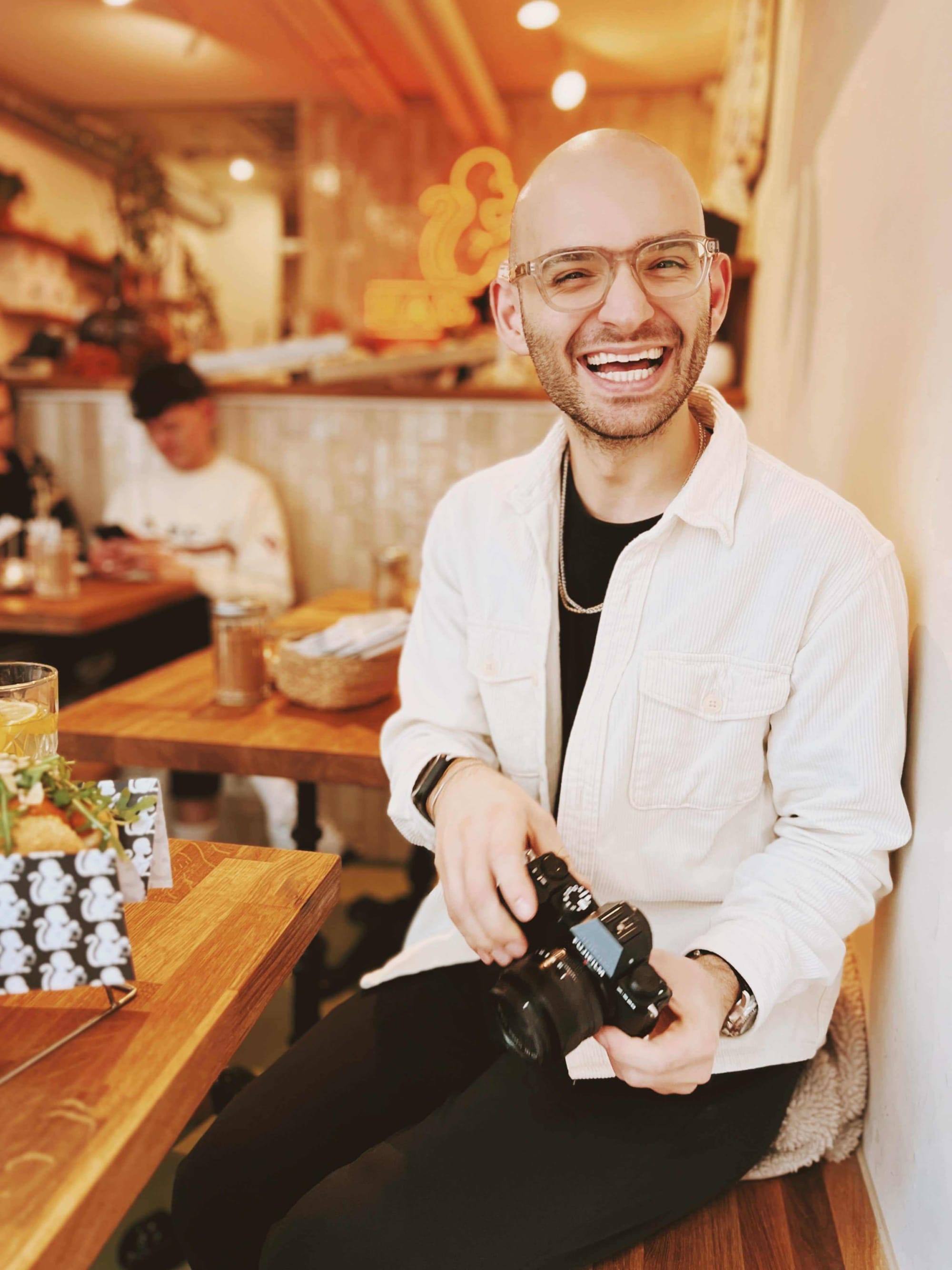 Glatzköpfiger Mann im Café mit Kamera, moderner Look, positives Lebensgefühl nach Haarausfall